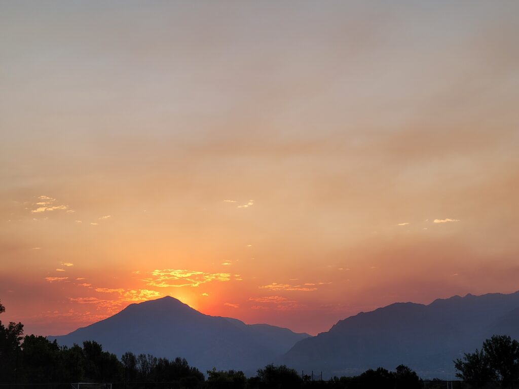 Sunrise glowing behind mountain silhouettes in Utah, with orange and pink skies softened by wildfire haze.
